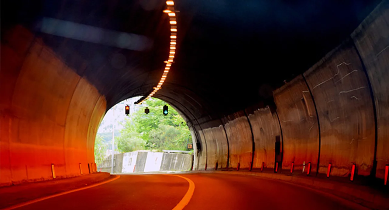 A curved road tunnel lit by warm orange ceiling lights, with a bright opening at the far end revealing green trees and daylight, and traffic signals hanging above the exit.